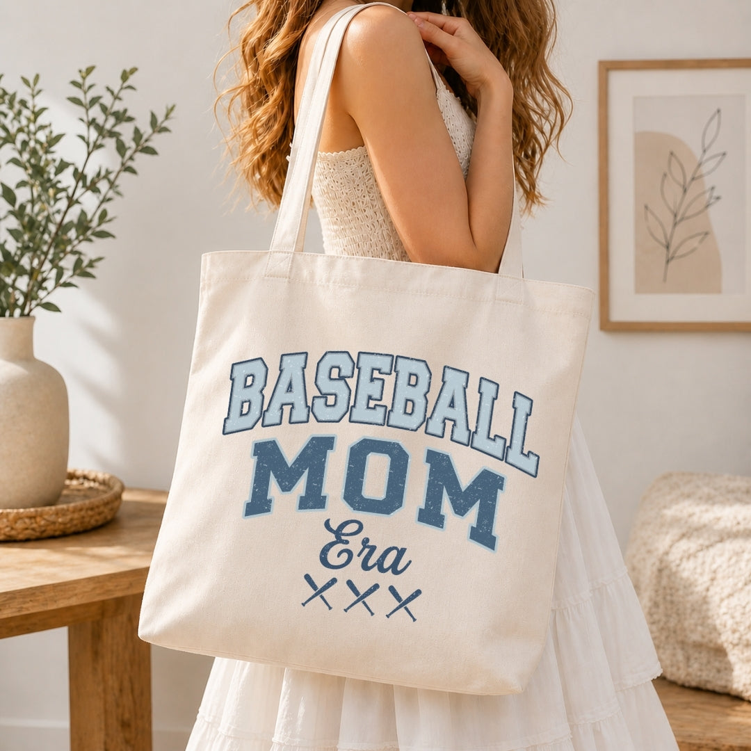 Woman in a white tiered dress carrying a canvas tote bag printed with a blue retro "Baseball Mom Era" graphic over her shoulder.