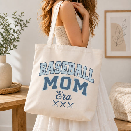 Woman in a white tiered dress carrying a canvas tote bag printed with a blue retro "Baseball Mom Era" graphic over her shoulder.