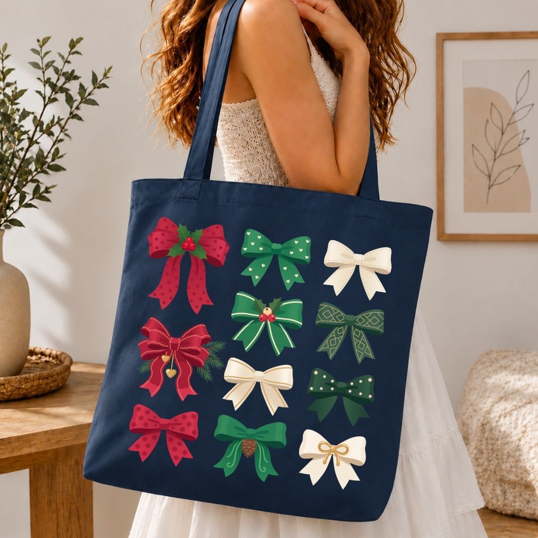 A navy blue canvas tote bag with a festive pattern of twelve Christmas bows in red, green, and white, carried by a woman in a white dress in a cozy indoor setting.