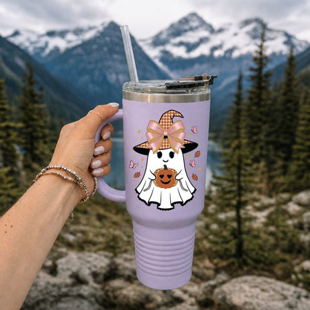 A hand holds a lavender insulated travel tumbler with a lid and straw, featuring a cartoon ghost wearing a witch hat and holding a pumpkin, set against a blurred mountain and forest background.