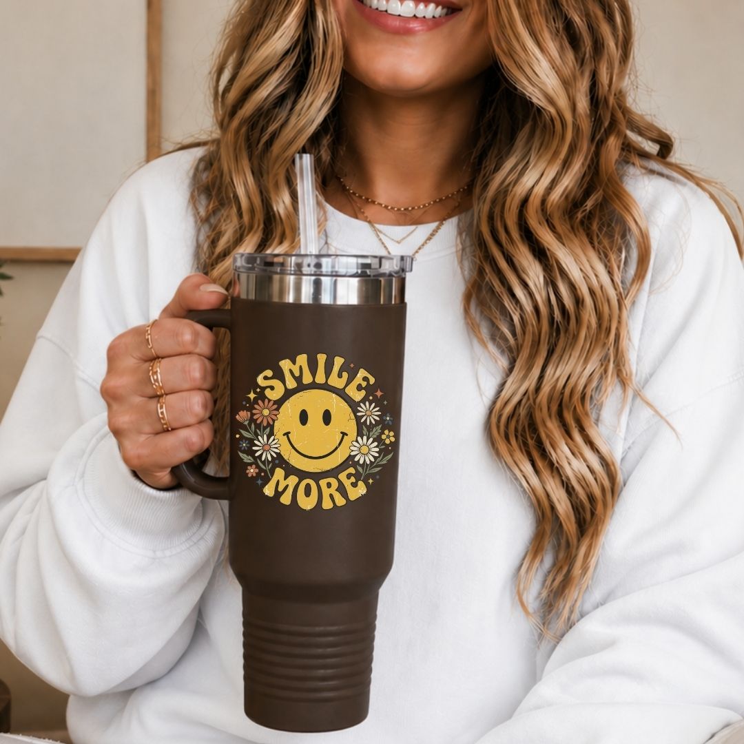 A woman holding a 40oz brown travel tumbler with a handle, featuring a vibrant sublimation print of a retro yellow smiley face design.