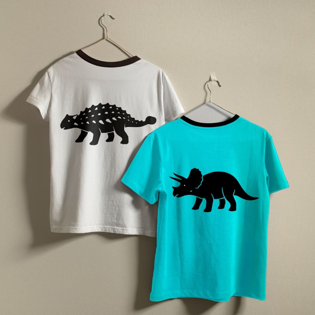 Two children's short-sleeve ringer T-shirts hanging on metal hangers against a beige wall. The left shirt is white with a black collar and a large black ankylosaurus graphic on the back.