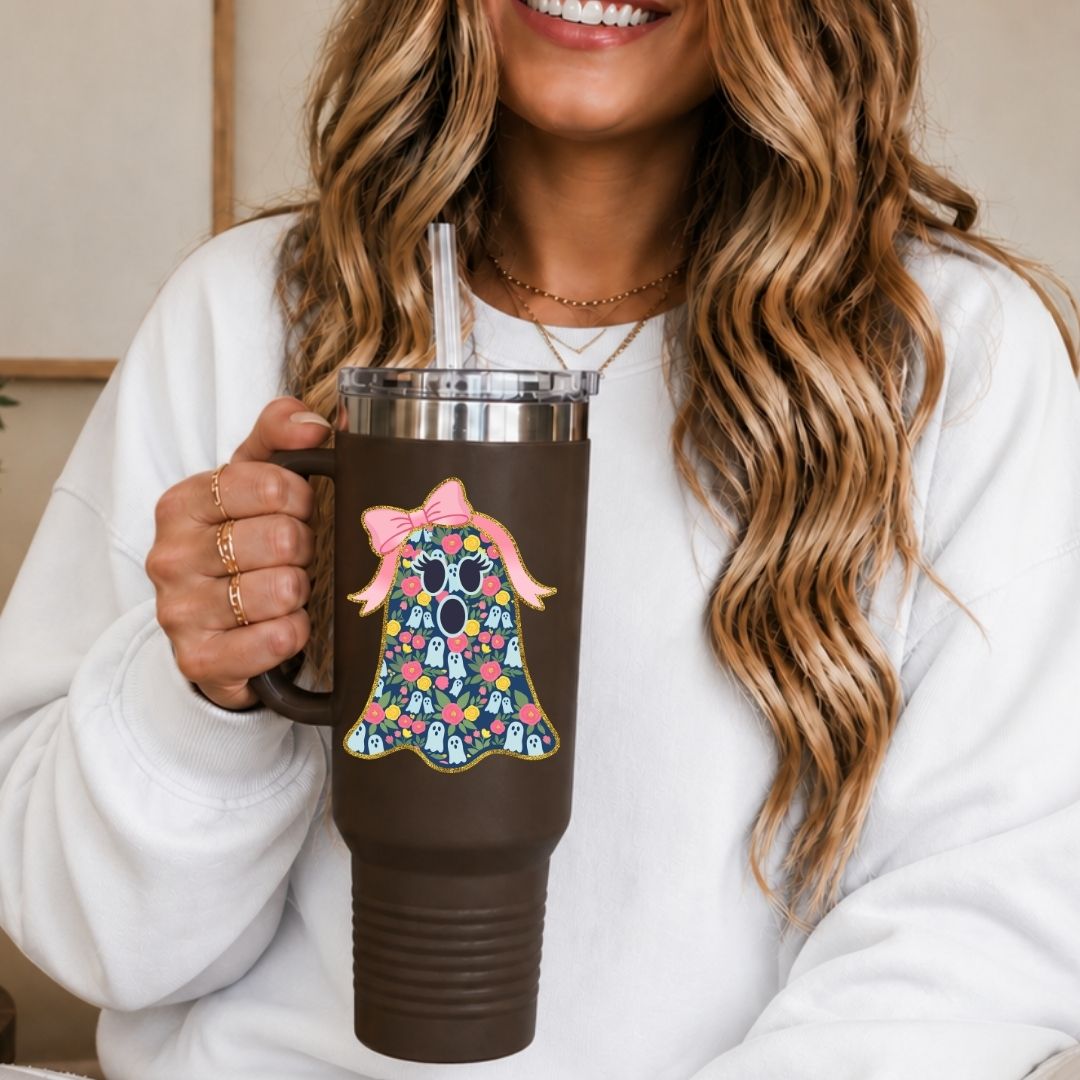A woman in a white sweatshirt holds a dark brown insulated tumbler with a clear lid and straw. The tumbler features a colorful graphic of a ghost with a pink bow and floral pattern.