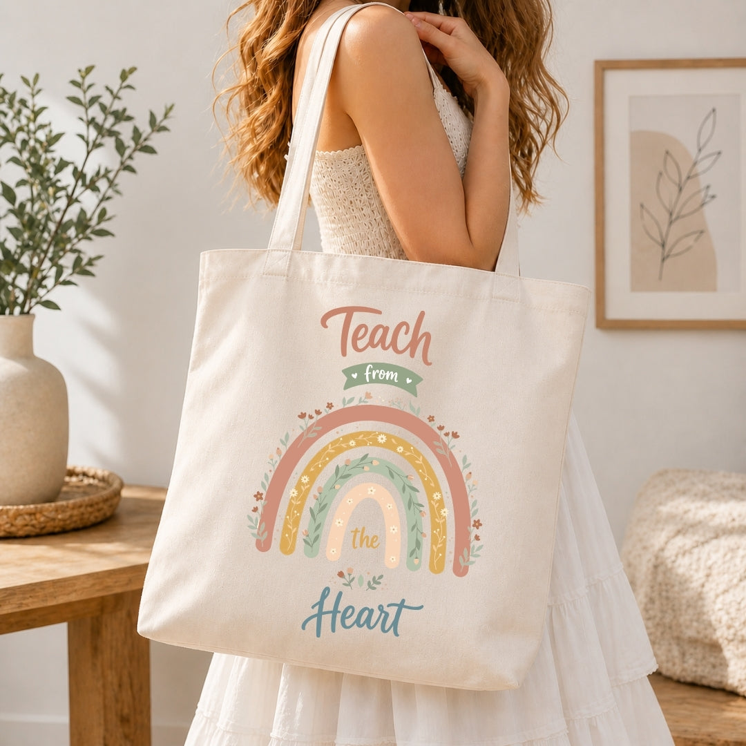 A woman in a white dress carries a cream canvas tote bag with a colorful rainbow and the words 'Teach from the Heart' printed on it, set in a bright, modern room.
