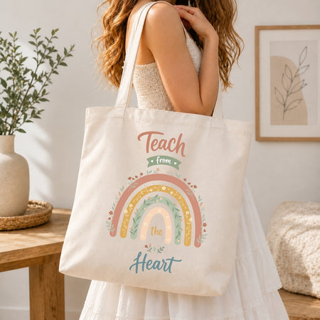 A woman in a white dress carries a cream canvas tote bag with a colorful rainbow and the words 'Teach from the Heart' printed on it, set in a bright, modern room.
