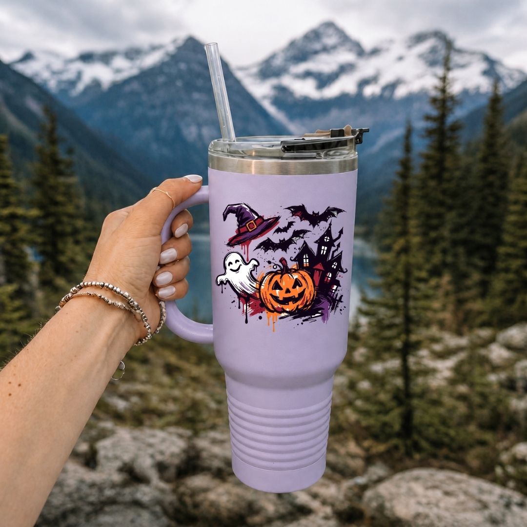 A lavender purple insulated travel tumbler with a clear straw, held by a hand against a mountain and forest background. The tumbler features a Halloween graphic with a jack-o'-lantern, ghost, witch hat, and bats.