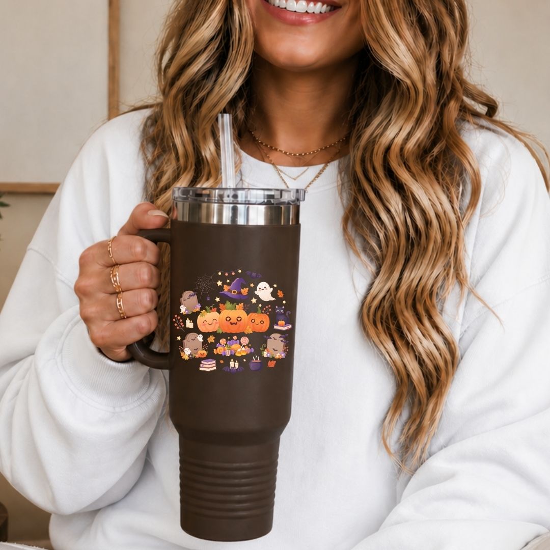 A woman in a white sweatshirt holds a brown insulated travel tumbler with a clear straw. The tumbler has a colorful Halloween-themed graphic featuring pumpkins, ghosts, and a witch on the front.