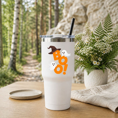 White travel tumbler with a black straw featuring an orange "BOO!" Halloween design, placed outdoors near a nature trail.