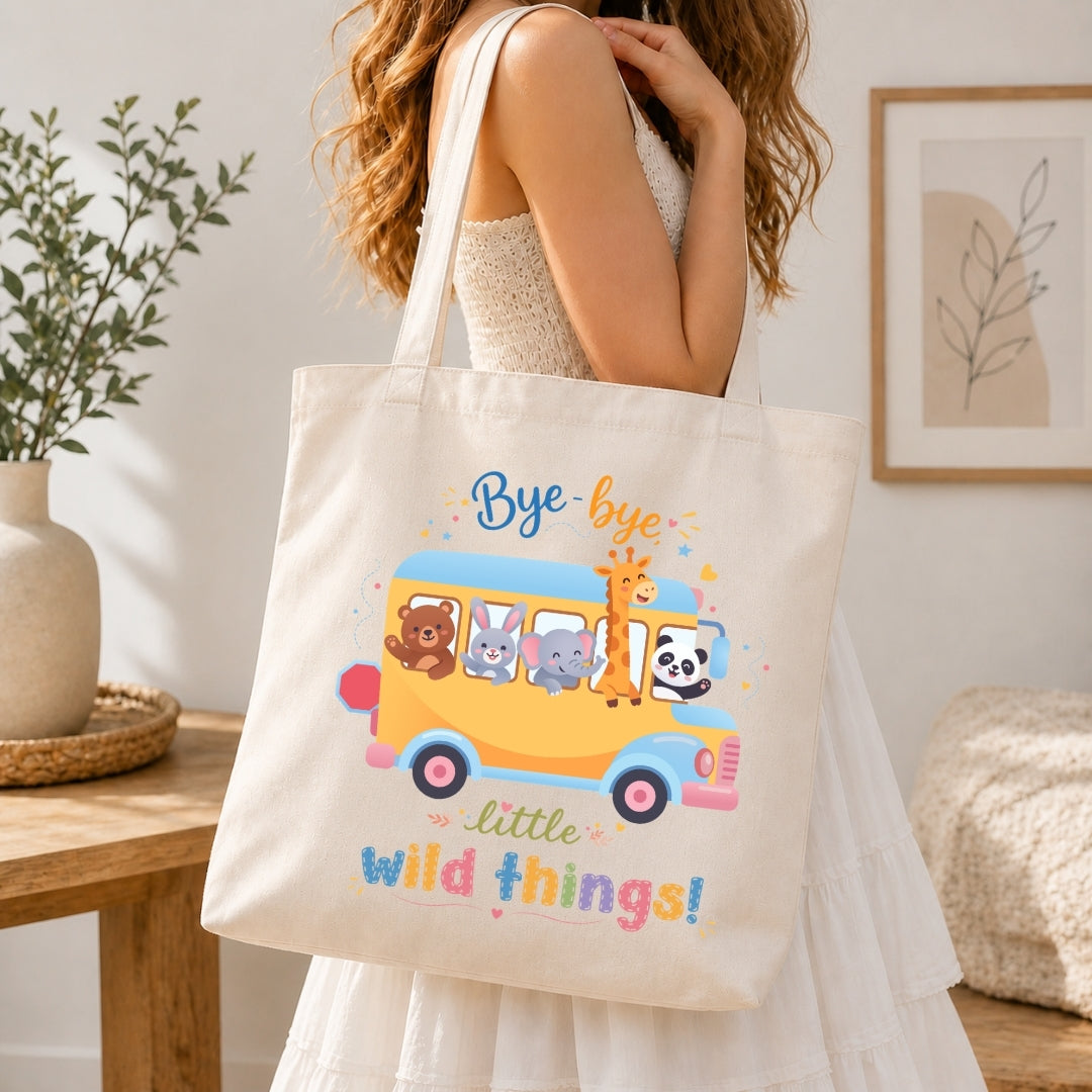 A person holding a cream-colored tote bag printed with a school bus graphic featuring cartoon animals and the text "Bye-bye little wild things!"