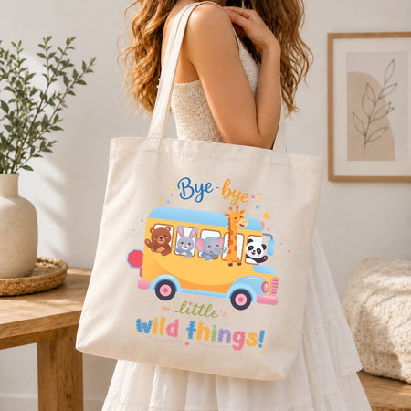 A person holding a cream-colored tote bag printed with a school bus graphic featuring cartoon animals and the text "Bye-bye little wild things!"