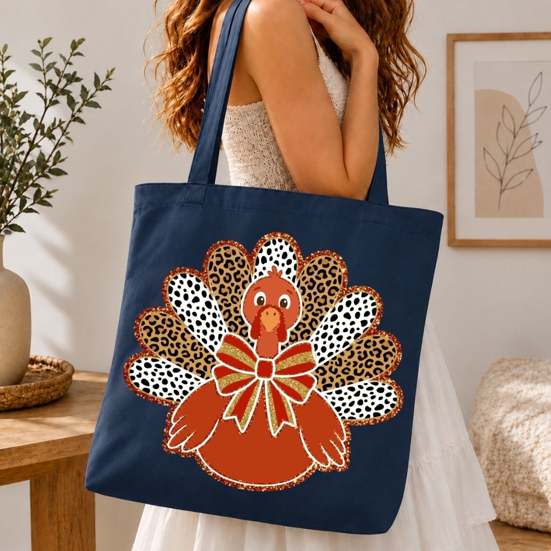 A navy blue canvas tote bag with a large autumn turkey graphic, carried by a woman in a white dress in a cozy indoor setting.