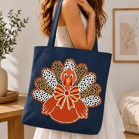 A navy blue canvas tote bag with a large autumn turkey graphic, carried by a woman in a white dress in a cozy indoor setting.
