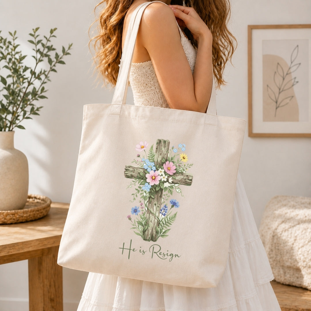 A woman holding a cream-colored tote bag printed with a floral cross graphic and the text "He is Resign".