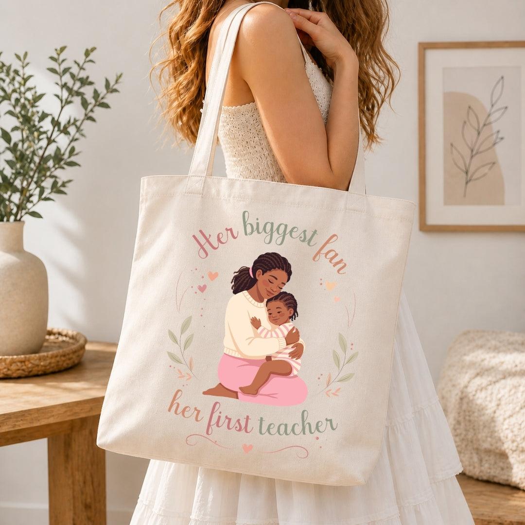 Woman carrying a cream-colored canvas tote bag over her shoulder featuring the mother-daughter graphic illustration and Her biggest fan, her first teacher text.