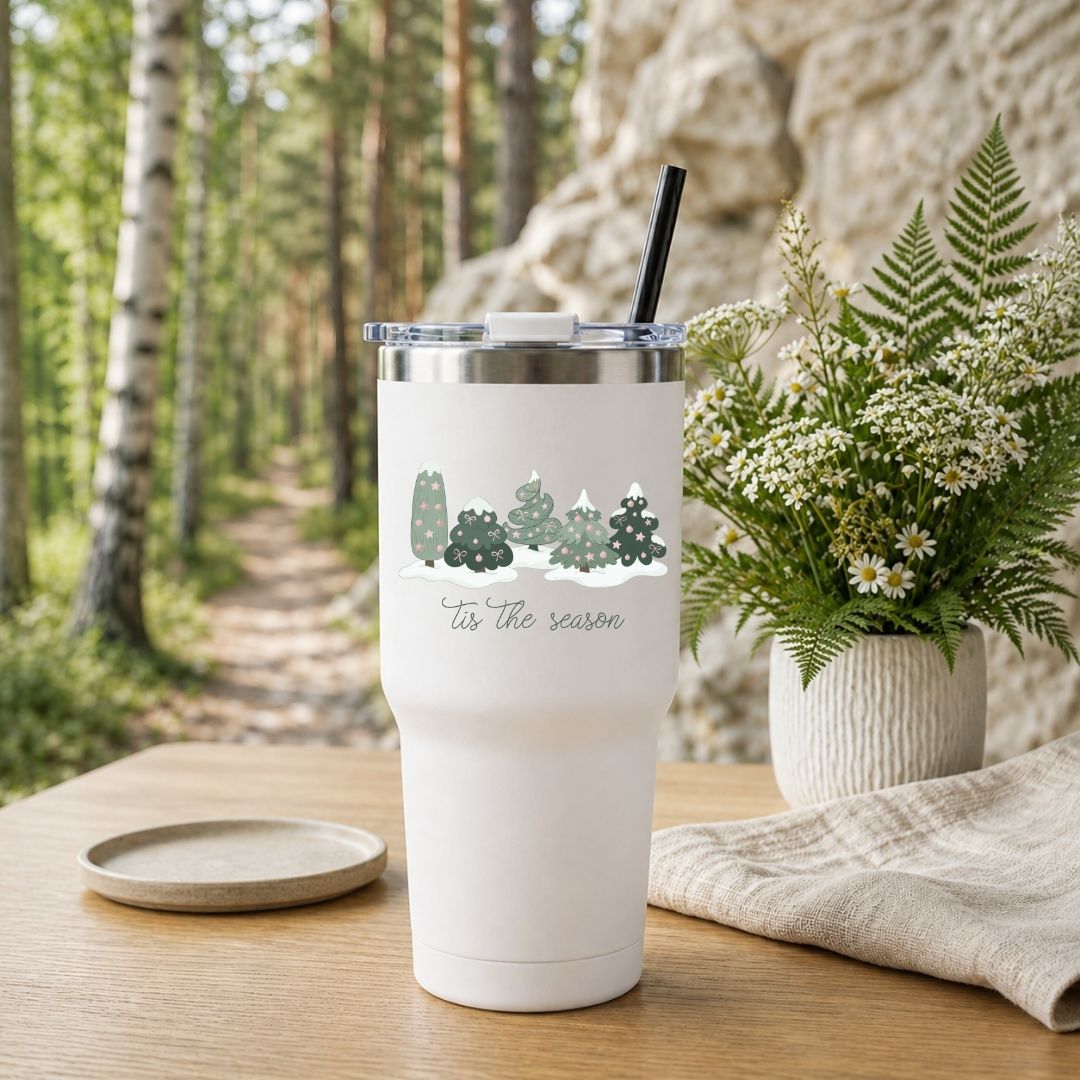 A white insulated tumbler with a clear lid and black straw sits on a wooden table outdoors. The tumbler has a printed cartoon graphic of sports figures and the text 'in my game day era.