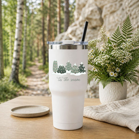 A white insulated tumbler with a clear lid and black straw sits on a wooden table outdoors. The tumbler has a printed cartoon graphic of sports figures and the text 'in my game day era.