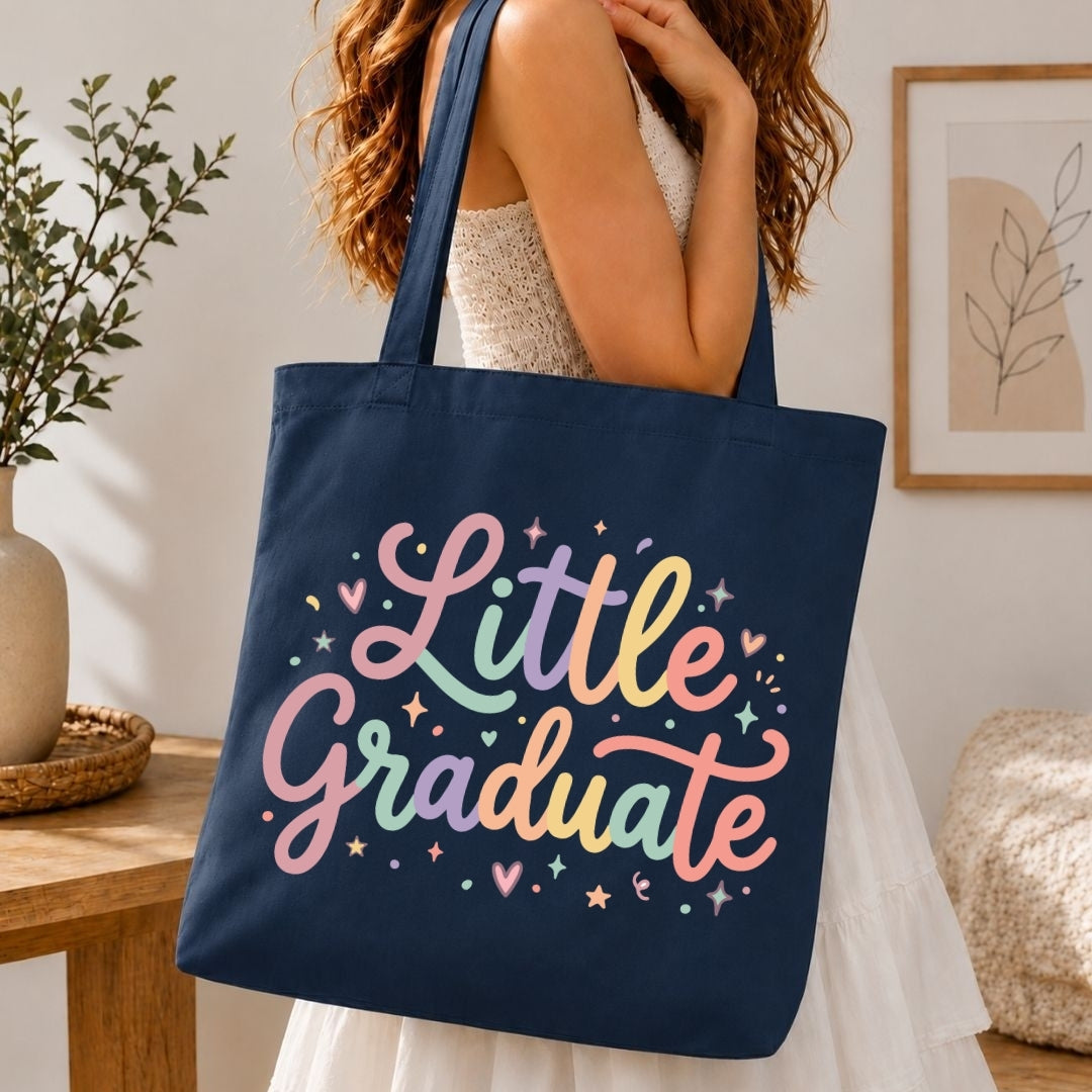 A woman wearing a white dress carries a navy blue canvas tote bag with the words 'Little Graduate' written in pastel rainbow script on the front, set in a bright, modern indoor room.