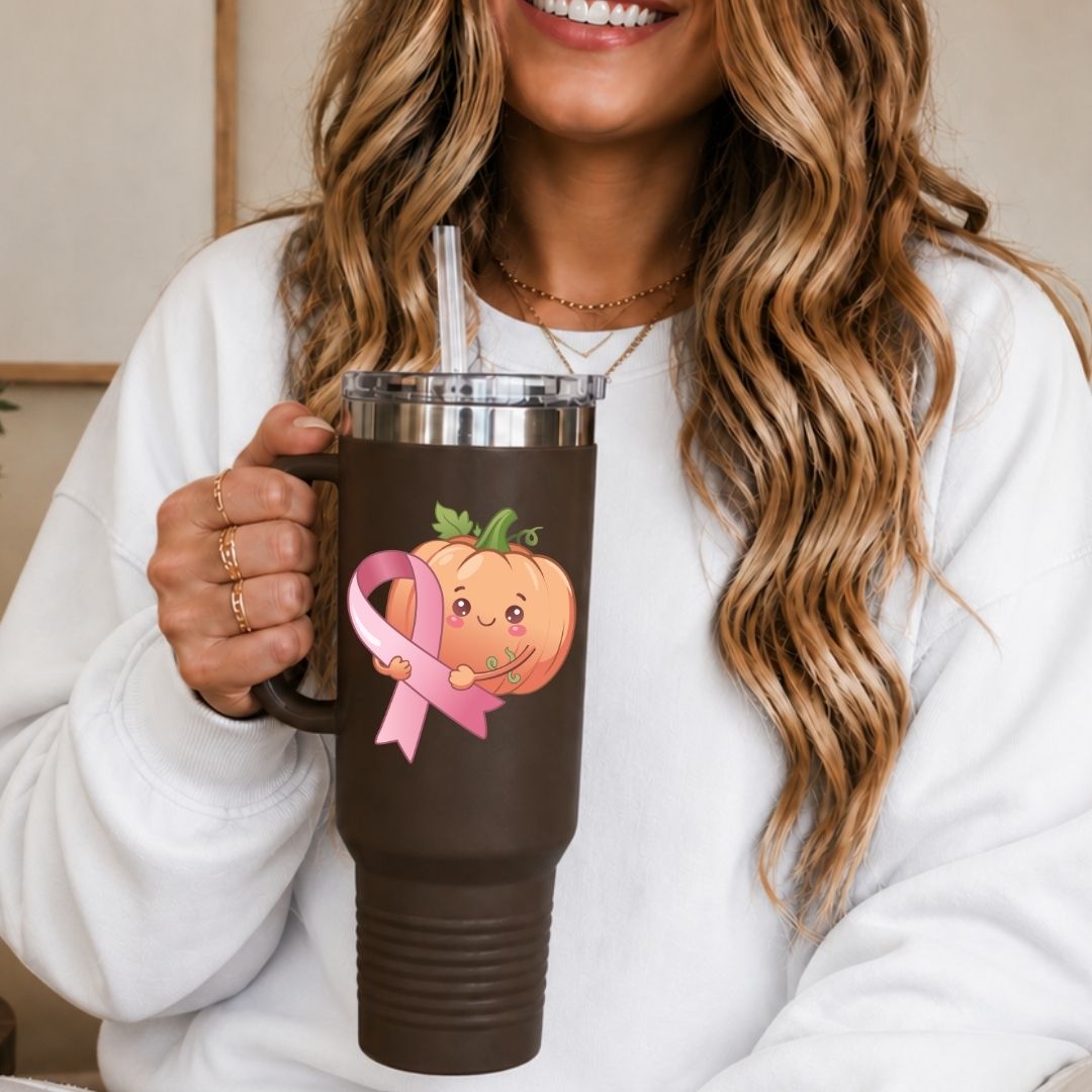 A woman in a white sweatshirt holds a dark brown insulated tumbler with a clear lid and straw. The tumbler features a cute graphic of a smiling orange pumpkin holding a pink awareness ribbon.