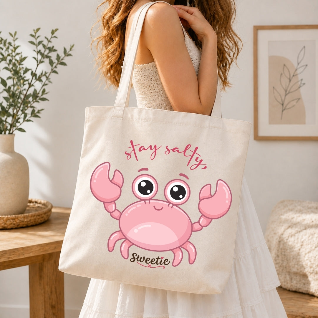 A woman holding a cream-colored tote bag printed with a cartoon pink crab graphic and the text "stay salty, Sweetie".