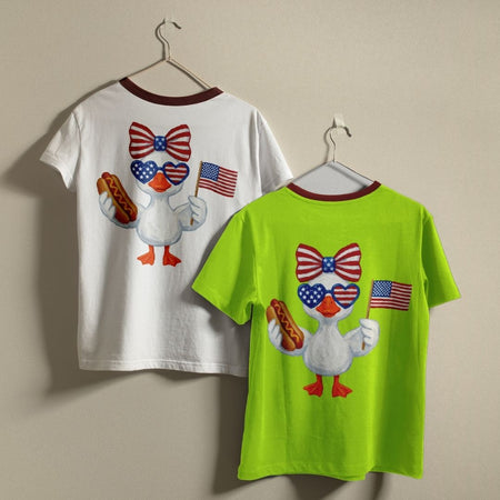 Two t-shirts, one white and one green, with a cartoon duck design on hangers against a beige background.