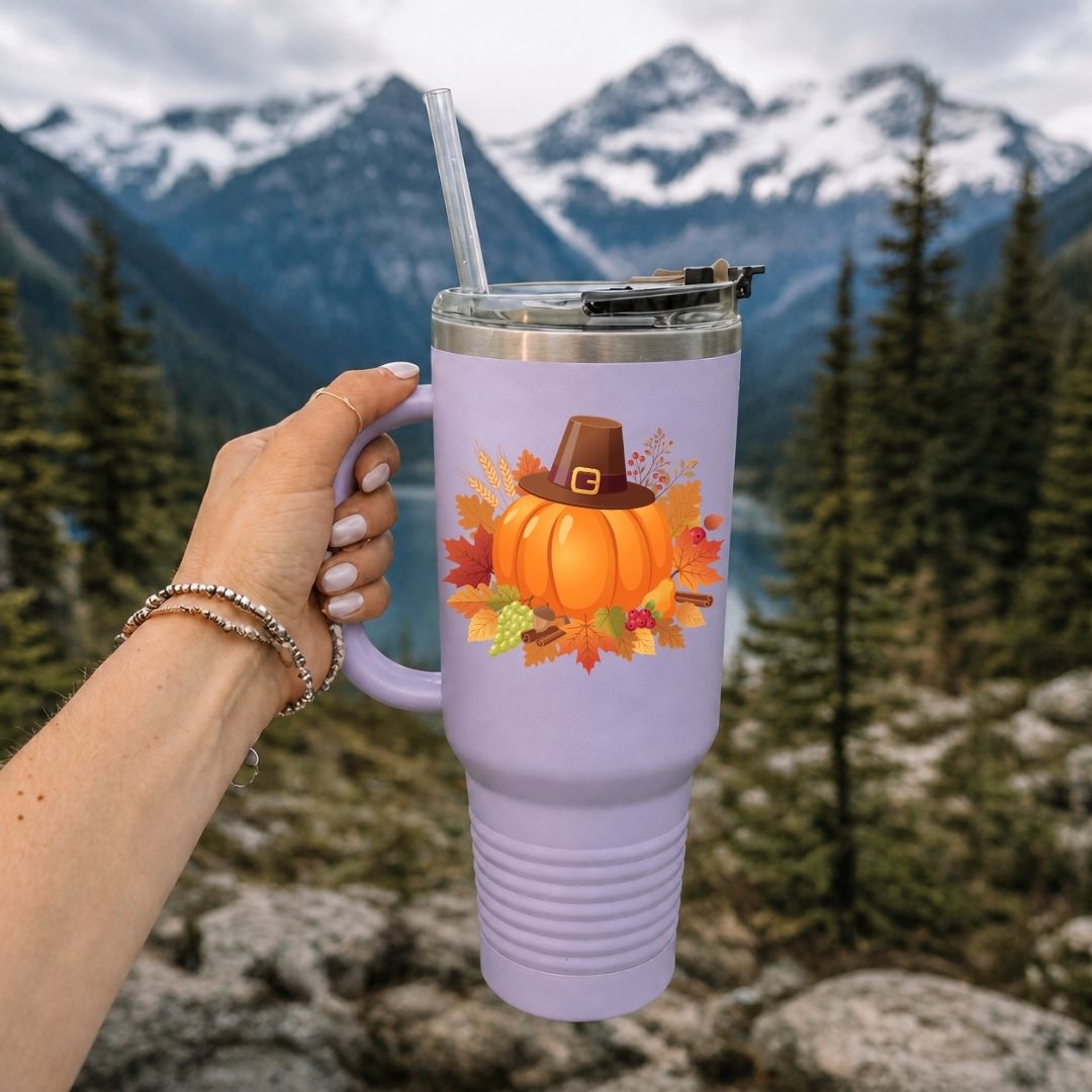 A lavender insulated tumbler with a handle, clear straw, and lid, held by a hand against a mountain and forest background. The tumbler features a graphic of an orange pumpkin wearing a brown pilgrim hat surrounded by autumn leaves.