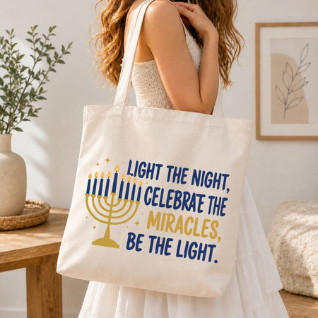 A woman wearing a white dress carries a cream canvas tote bag with a Hanukkah design. The bag features a gold menorah and the text 'LIGHT THE NIGHT, CELEBRATE THE MIRACLES, BE THE LIGHT.