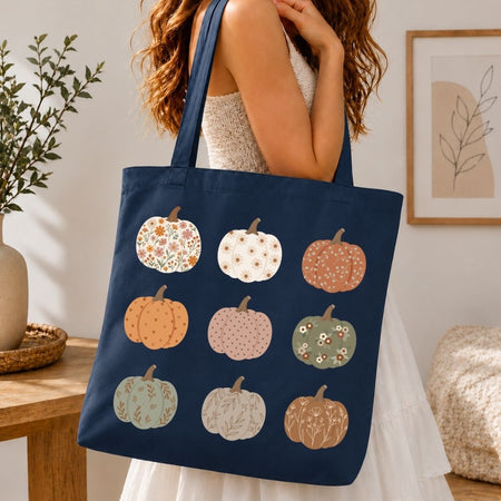 A navy blue canvas tote bag with a pattern of nine illustrated pumpkins in autumn colors, carried on the shoulder of a woman in a white dress in a warm, cozy room setting.