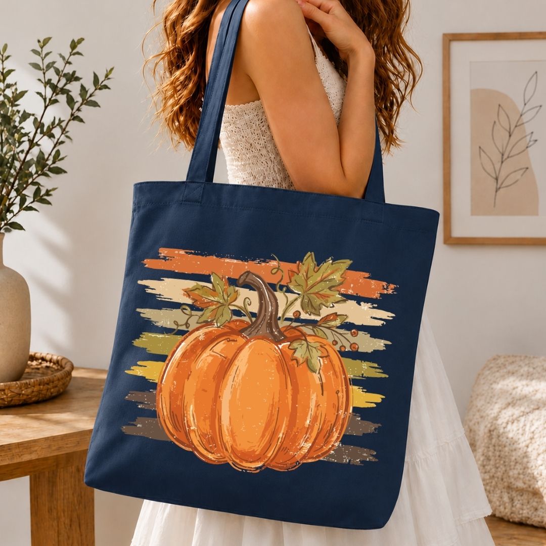 A woman wearing a white dress carries a navy blue canvas tote bag with a large orange pumpkin and autumn leaf graphic on the front, set in a warm, cozy indoor room.