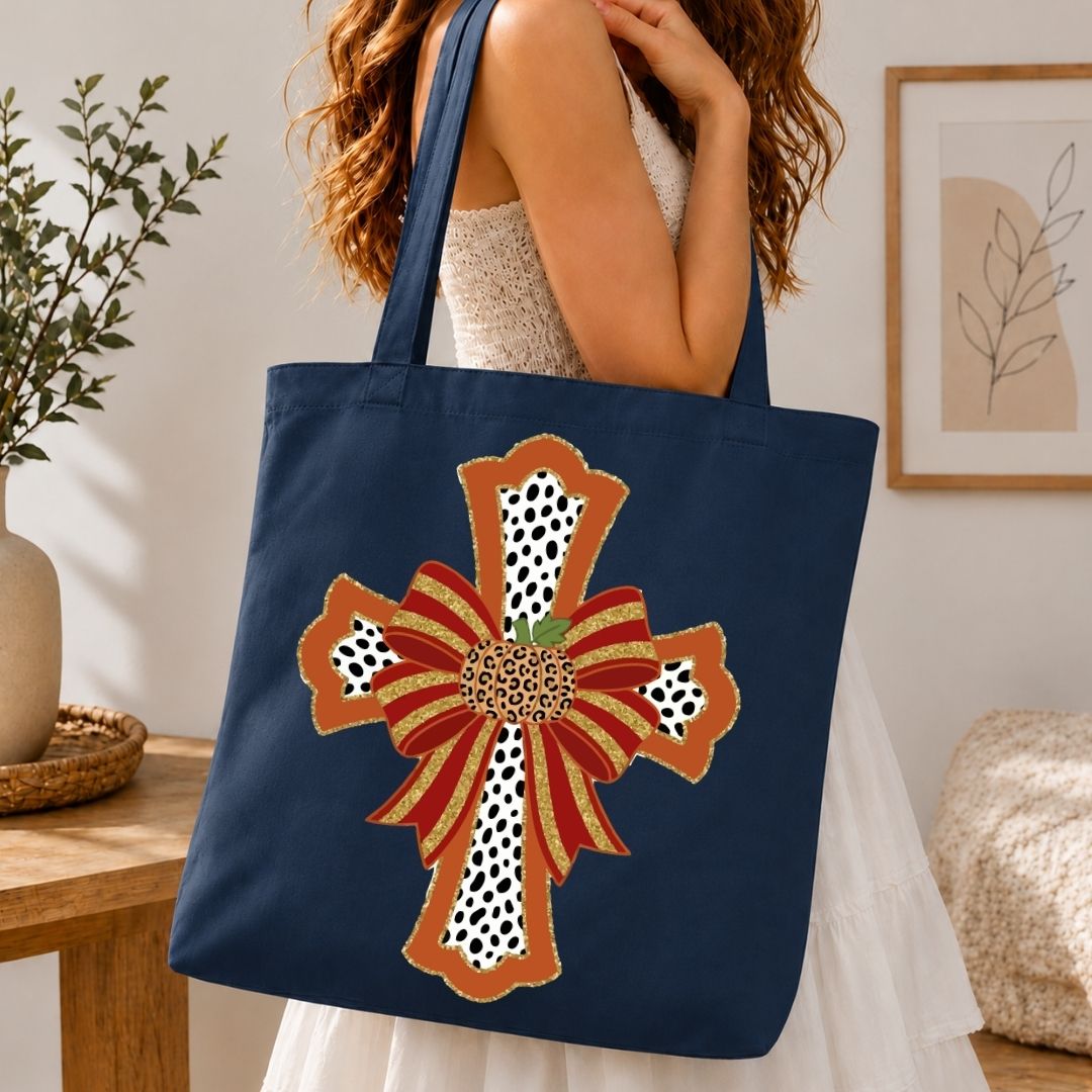 A navy blue canvas tote bag with a large decorative cross design in rust orange, red, white, and black, carried by a woman in a white dress in a warm, modern room.