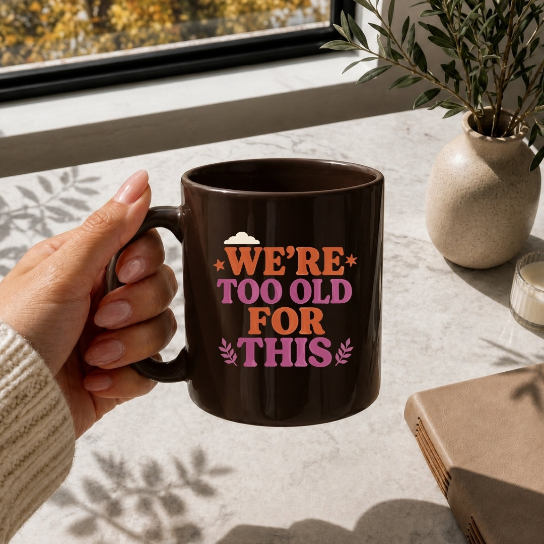 Person holding a mug with 'We're too old for this' text on a table with a window and plant in the background