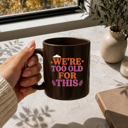 Person holding a mug with 'We're too old for this' text on a table with a window and plant in the background