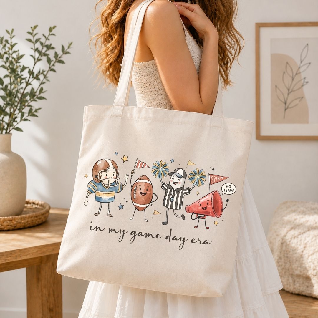 A woman in a white dress carries a cream canvas tote bag with a colorful cartoon graphic of sports figures and the text 'in my game day era'. The scene is set in a bright, modern room with a wooden table, potted plant, and framed art.