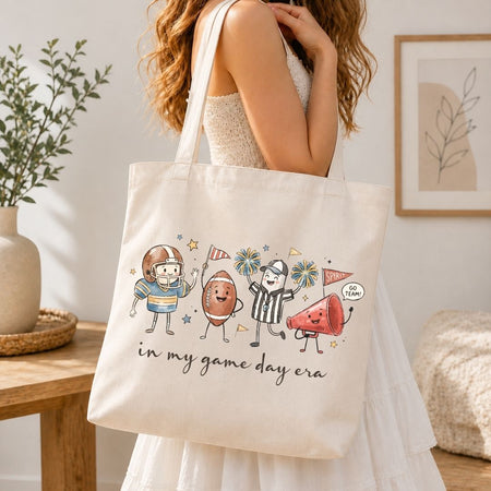 A woman in a white dress carries a cream canvas tote bag with a colorful cartoon graphic of sports figures and the text 'in my game day era'. The scene is set in a bright, modern room with a wooden table, potted plant, and framed art.