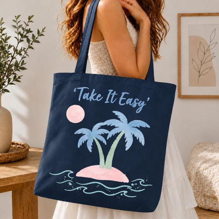 A woman wearing a white dress carries a navy blue canvas tote bag with a tropical beach design. The bag features the text 'Take It Easy' in light blue script, above two palm trees on a pink sand island with light blue waves and a pink sun.