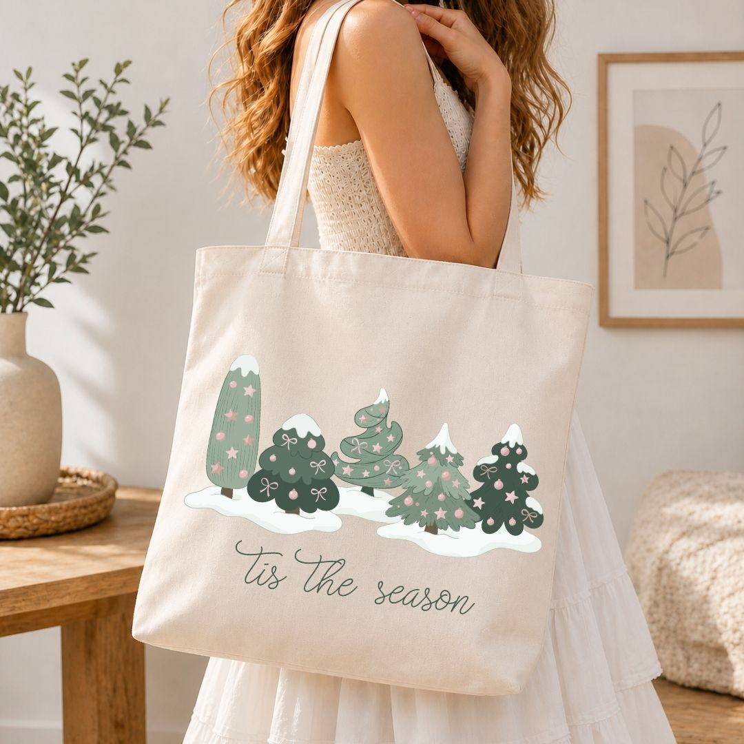 A woman in a white dress carries a cream canvas tote bag with a winter tree illustration and the text 'Tis the season'.