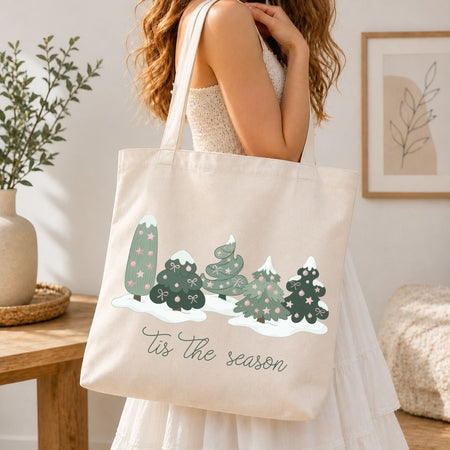A woman in a white dress carries a cream canvas tote bag with a winter tree illustration and the text 'Tis the season'.