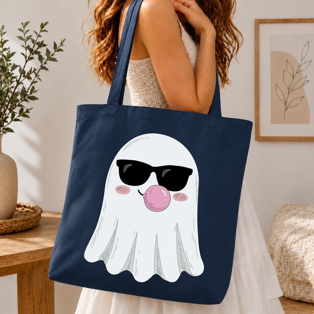A navy blue canvas tote bag with a cartoon ghost graphic wearing black sunglasses and blowing a pink bubblegum bubble, carried by a woman in a white dress in a cozy indoor setting.