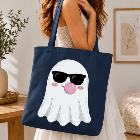 A navy blue canvas tote bag with a cartoon ghost graphic wearing black sunglasses and blowing a pink bubblegum bubble, carried by a woman in a white dress in a cozy indoor setting.