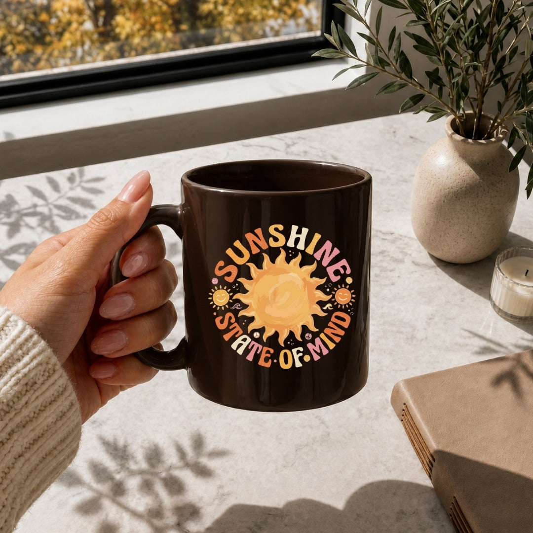 Person holding a mug with 'Sunshine State of Mind' design on a table with a window and plant in the background.