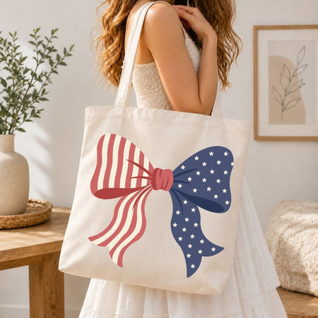 A woman carrying a cream-colored canvas tote bag over her shoulder printed with a large, vibrant American flag coquette ribbon bow design.