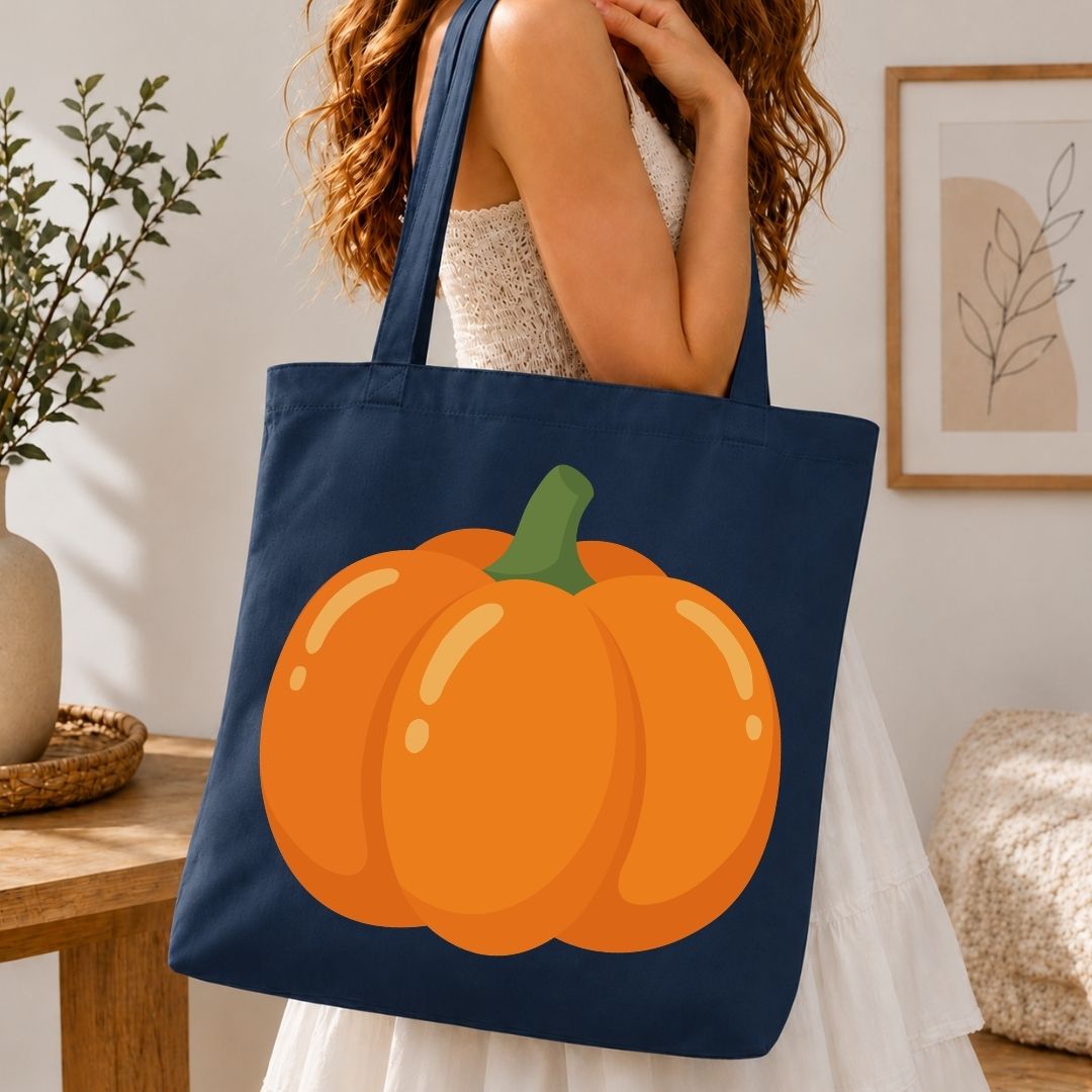 A navy blue canvas tote bag with a large orange pumpkin graphic on the front, carried by a woman in a white dress in a cozy indoor setting.