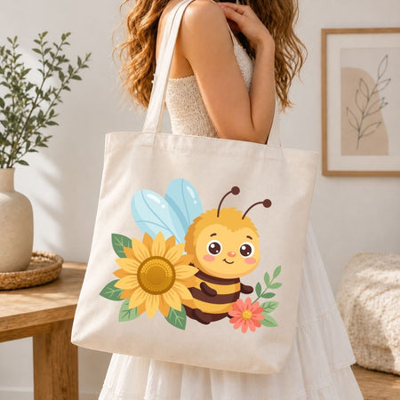A woman wearing a white dress carries a cream canvas tote bag decorated with a cute cartoon bee, yellow sunflower, and pink flower. The scene is set in a bright, modern room with a wooden table, potted plant, and framed wall art.
