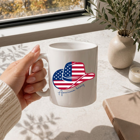 White mug with American flag design held by a hand on a table with a plant and notebook.