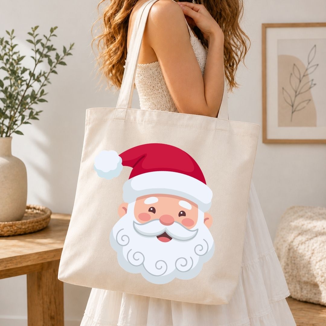 A woman wearing a white dress carries a cream canvas tote bag featuring a cute cartoon Santa Claus face graphic. The setting is a bright, modern room with a wooden table, potted plant, and framed wall art.