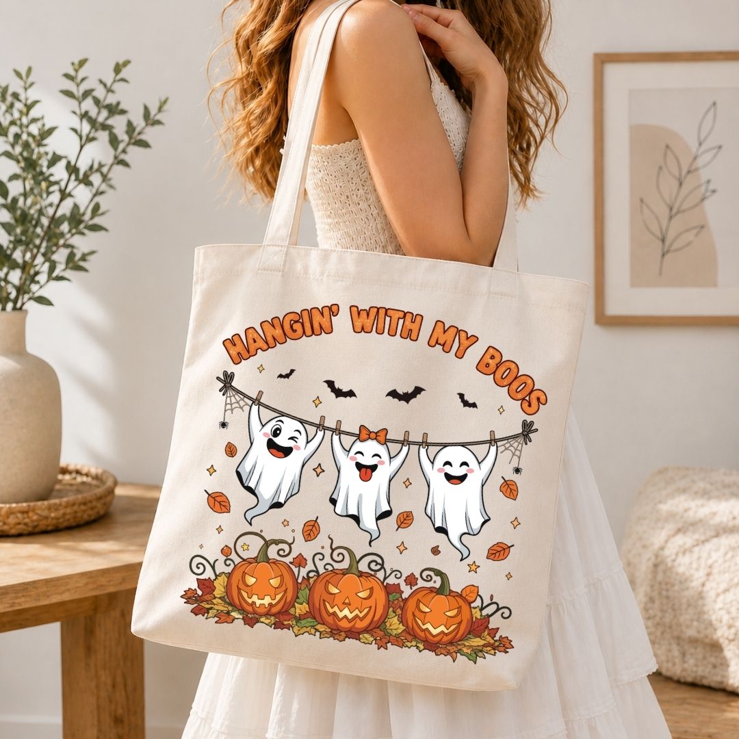 A woman in a white dress carrying a natural canvas tote bag printed with three cute ghosts hanging from a clothesline above pumpkins and fall leaves.