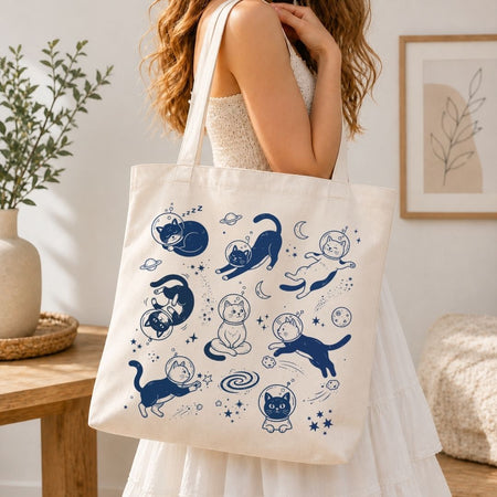 A woman in a white dress carries a cream canvas tote bag decorated with blue cartoon cat illustrations in space, set in a bright, modern room.