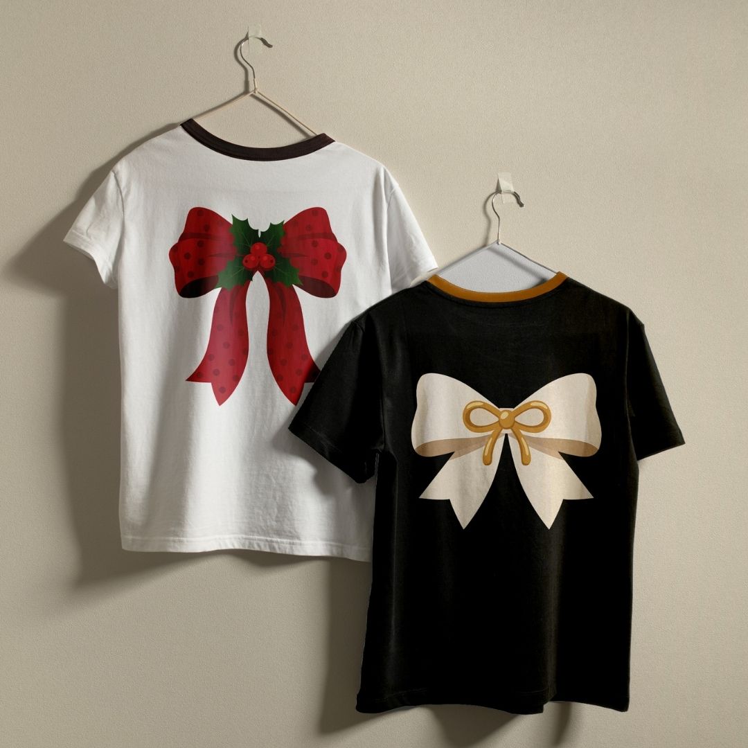 Two short-sleeve graphic T-shirts hanging on hangers against a plain beige wall. The left shirt is white with a dark collar trim and a large red bow graphic with green holly leaves on the back.
