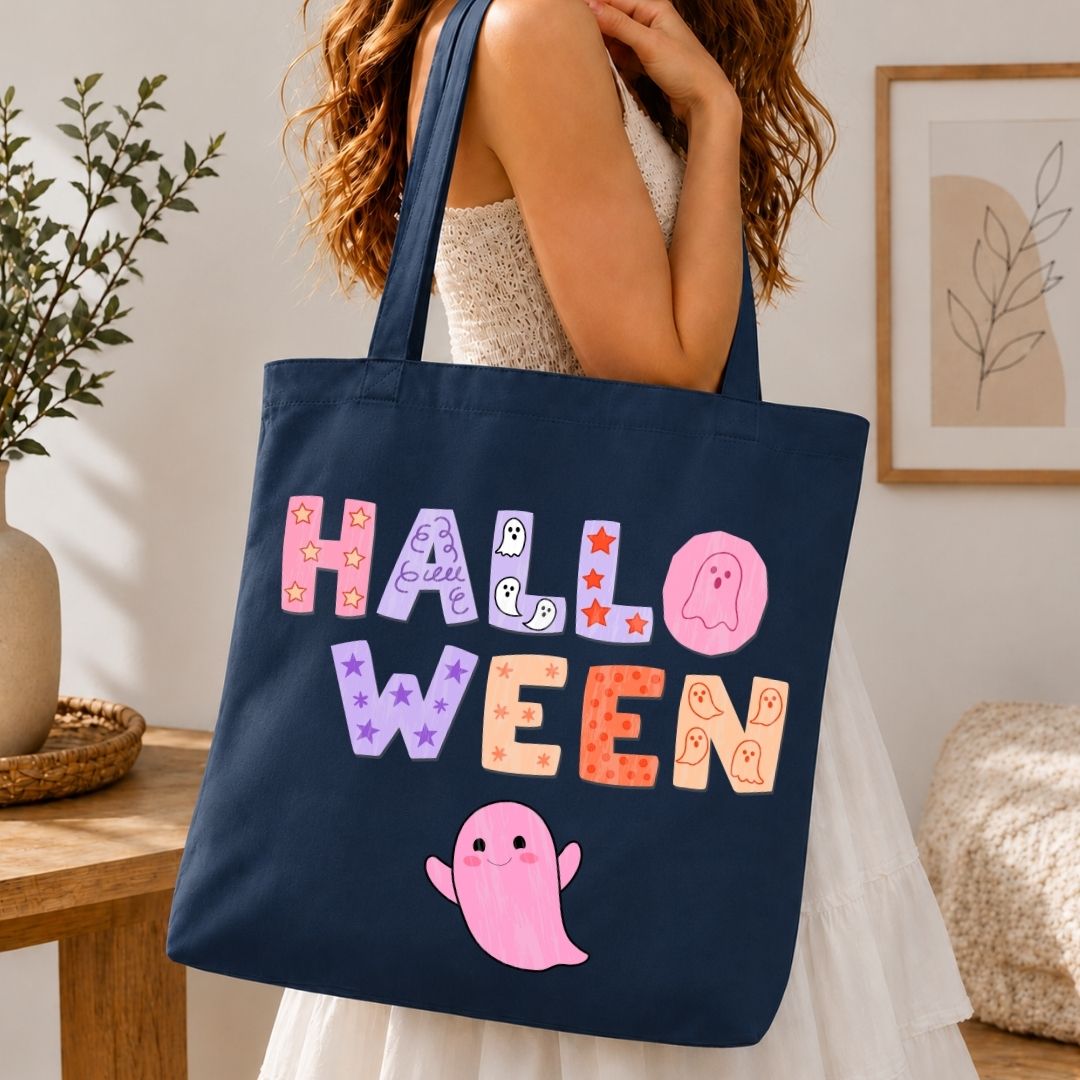 A navy blue canvas tote bag with a colorful 'HALLOWEEN' graphic and a pink ghost, carried by a woman in a white dress in a cozy indoor setting.