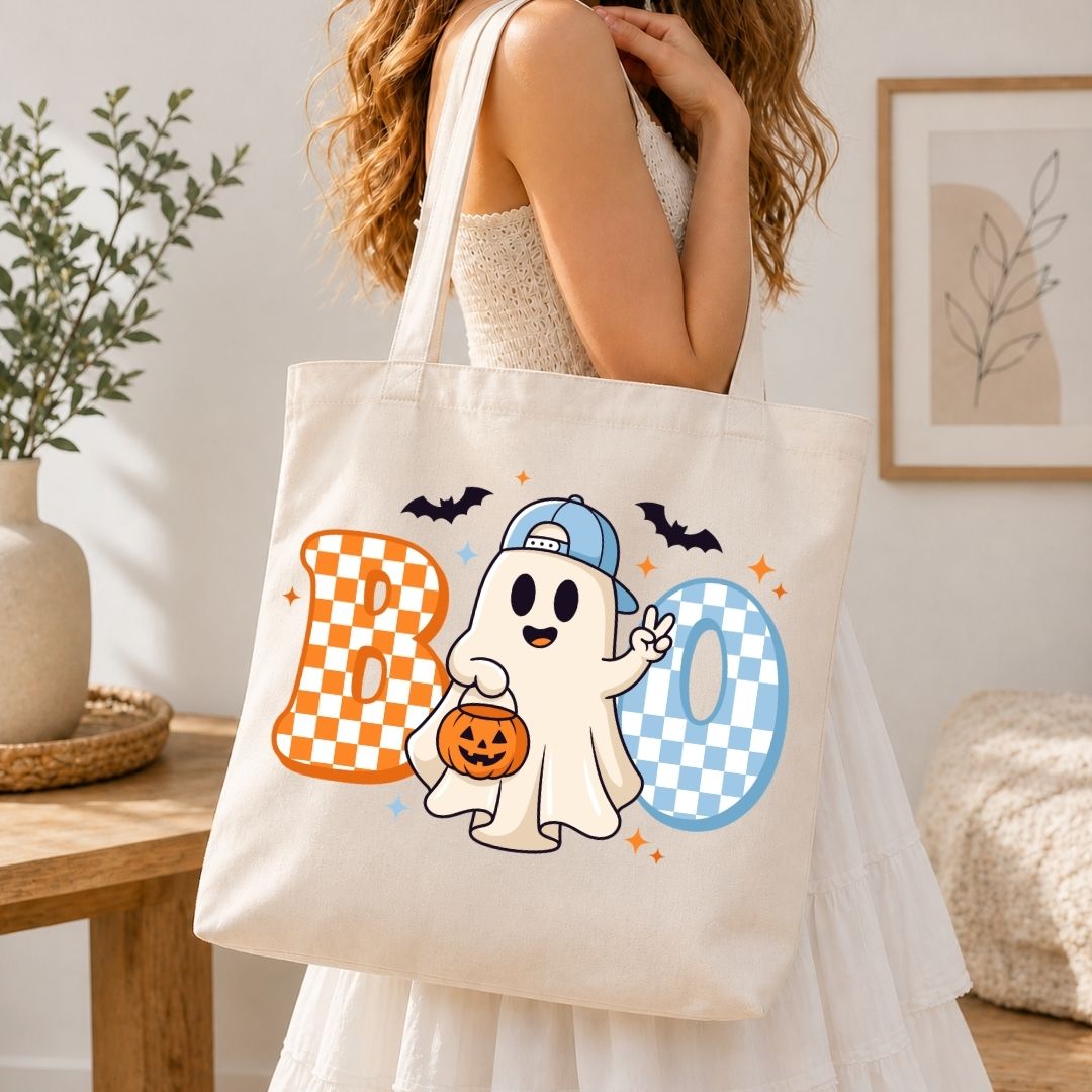 Woman holding a canvas tote bag printed with a cute Halloween ghost wearing a cap and checkered "BOO" text design.