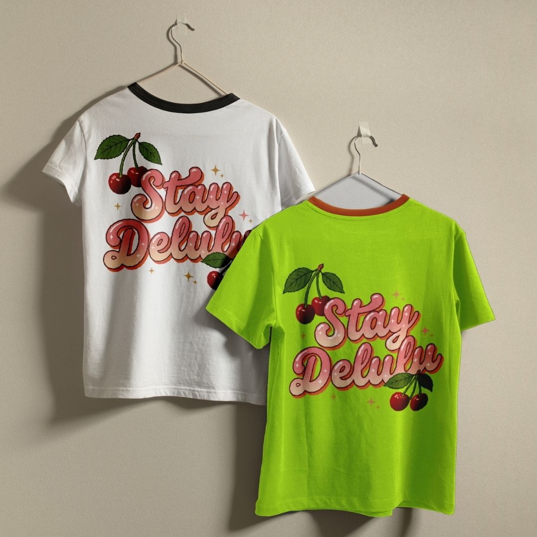 Two t-shirts, one white and one green, with 'Stay Delux' text and cherry graphics on hangers against a beige background.